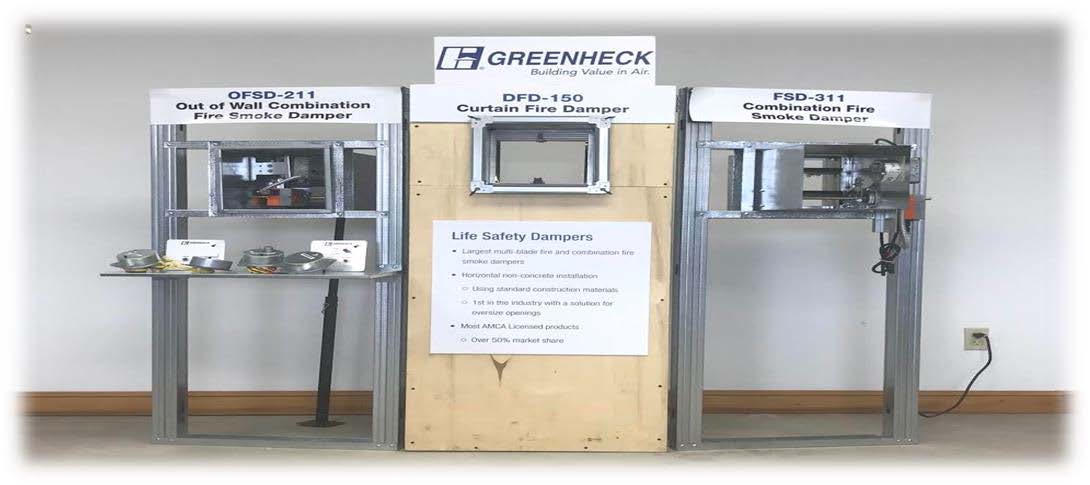 Display of three Greenheck damper models with labels: OFSD-211 Out of Wall Combination Fire Smoke Damper, DFD-150 Curtain Fire Damper, and FSD-311 Combination Fire Smoke Damper. Includes a description of life safety dampers on a central wooden panel and the company's logo above.