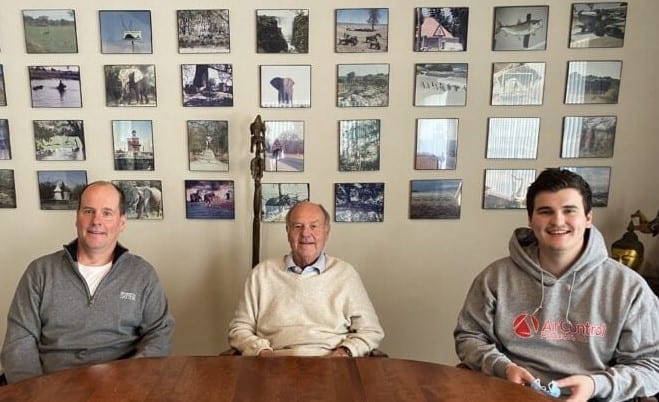 Three Men Sitting In Front Of Photo Wall | Air Control Products Three men sitting at a wooden table with a wall of photographs behind them. The man on the left is wearing a gray jacket, the man in the middle is wearing a white sweater, and the man on the right is wearing a gray hoodie. The photographs on the wall are arranged in a grid pattern and depict various scenes and objects.