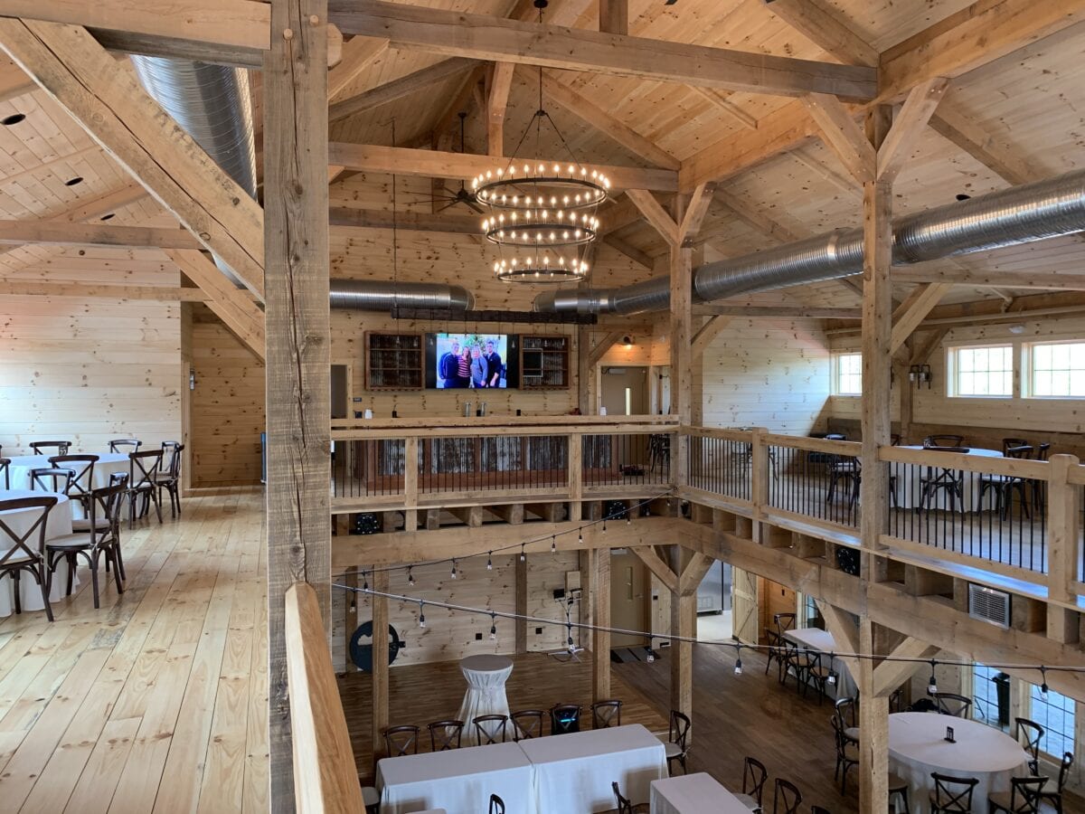 Interior view of a rustic barn-style event space with wooden beams and floors. The upper level features seating with round and square tables and cross-back chairs. A large, circular chandelier is suspended from the ceiling. Visible ductwork runs along the ceiling, and a flat-screen TV is mounted on the wall.