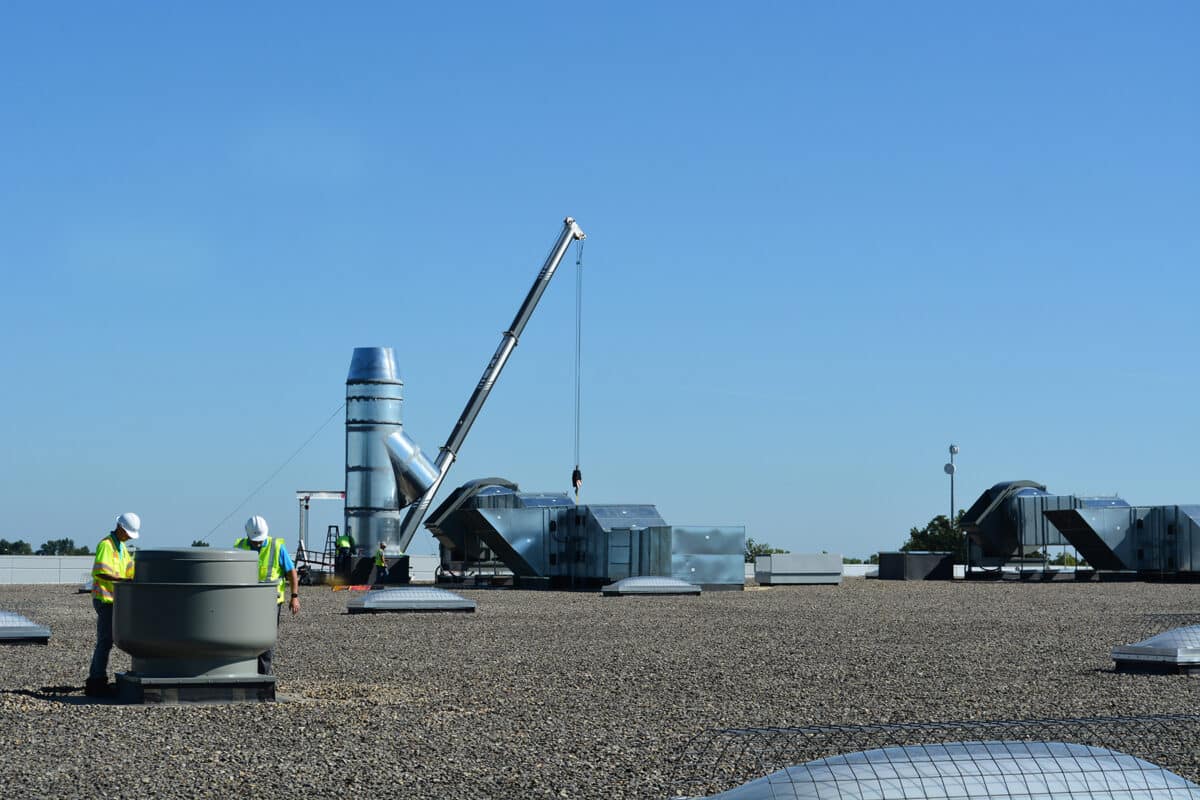 Two workers in safety gear on a flat gravel rooftop working on HVAC equipment. A crane lifts a metal component into position near large ductwork and ventilation systems under a clear blue sky.