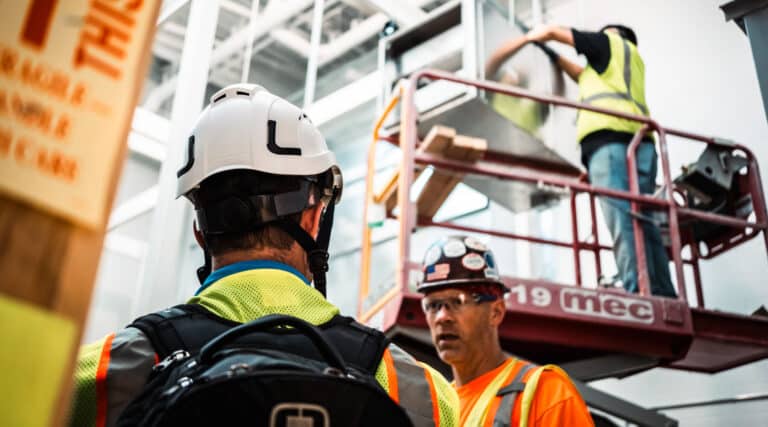 Construction workers on site; one wearing a hard hat and a safety vest is seen from the back, another wearing a hard hat with stickers and glasses is in conversation, and a third worker in a safety vest is elevated on a cherry picker working on metal ductwork.