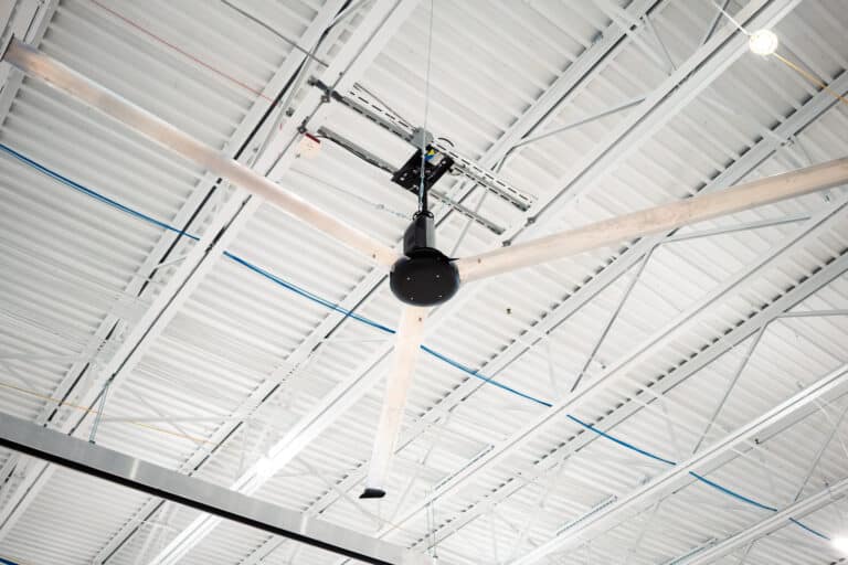A large industrial ceiling fan with multiple blades suspended from the ceiling of a warehouse. The ceiling is made of corrugated metal panels with exposed beams and various electrical cables running across it.