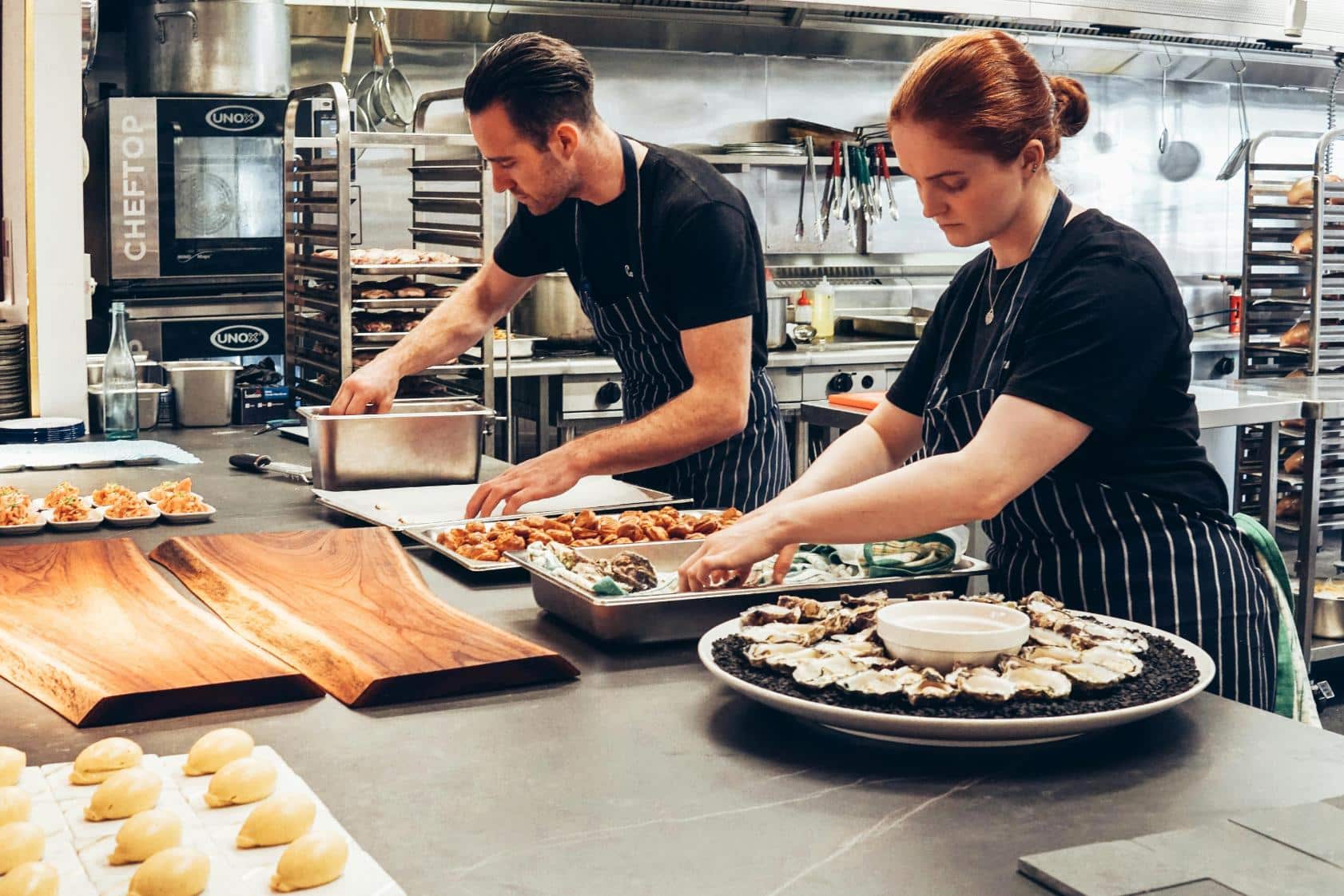 Two chefs wearing aprons are working in a commercial kitchen. One is placing food into trays while the other is arranging items on a large platter. The kitchen is equipped with industrial ovens and various cooking utensils. Several prepared dishes are visible on the countertop.