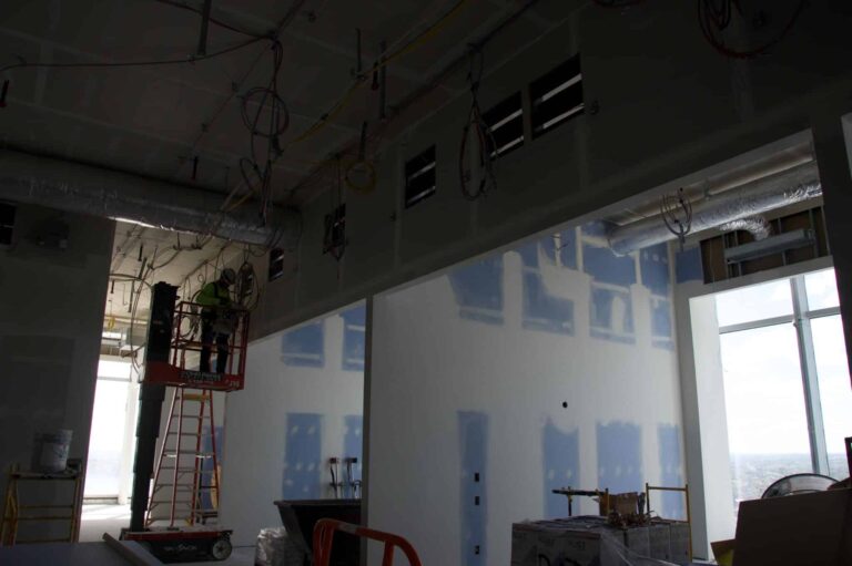 Interior construction site showing a partially built room with exposed ducts, wires, and unfinished walls. A worker on a lift is adjusting equipment near the ceiling. Sunlight streams in through large windows illuminating the work area.