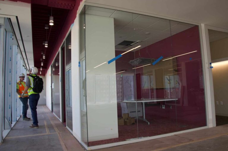 A corridor in a construction site with two workers wearing safety vests and hard hats standing near a glass wall. The corridor has unfinished flooring and overhead lights, with transparent glass panels on one side showing an interior room with unfinished ceiling and construction materials.