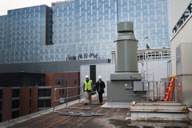 Two construction workers wearing safety vests and helmets on a rooftop with industrial equipment in the foreground, with a large modern building in the background.