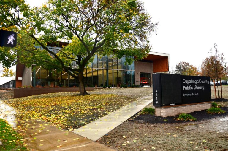 Exterior of Cuyahoga County Public Library's Brooklyn Branch, featuring a modern glass and brick building surrounded by trees with autumn leaves on the ground. A large sign displays the library's name and address, 7619.