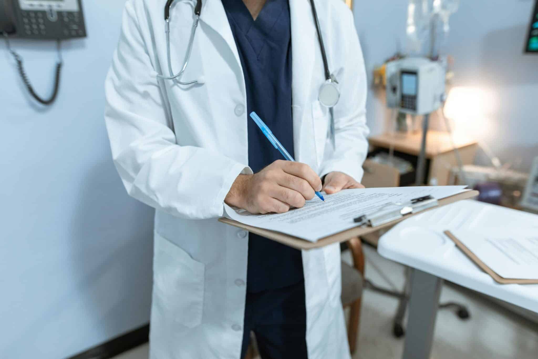 A doctor in a white coat is writing with a blue pen on a clipboard. The doctor is in a medical setting with a stethoscope around their neck and medical equipment visible in the background.