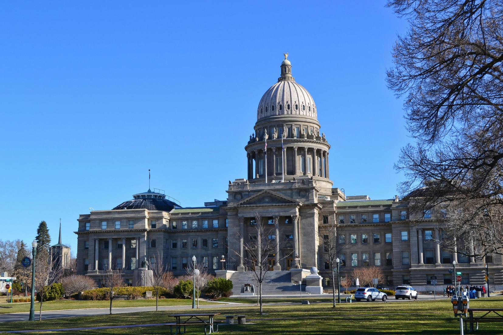 View of the Idaho State Capitol Building with its large dome and classical architecture, surrounded by a clear blue sky and trees. The grand entrance is framed by columns and flags, and cars and people are visible in the foreground.