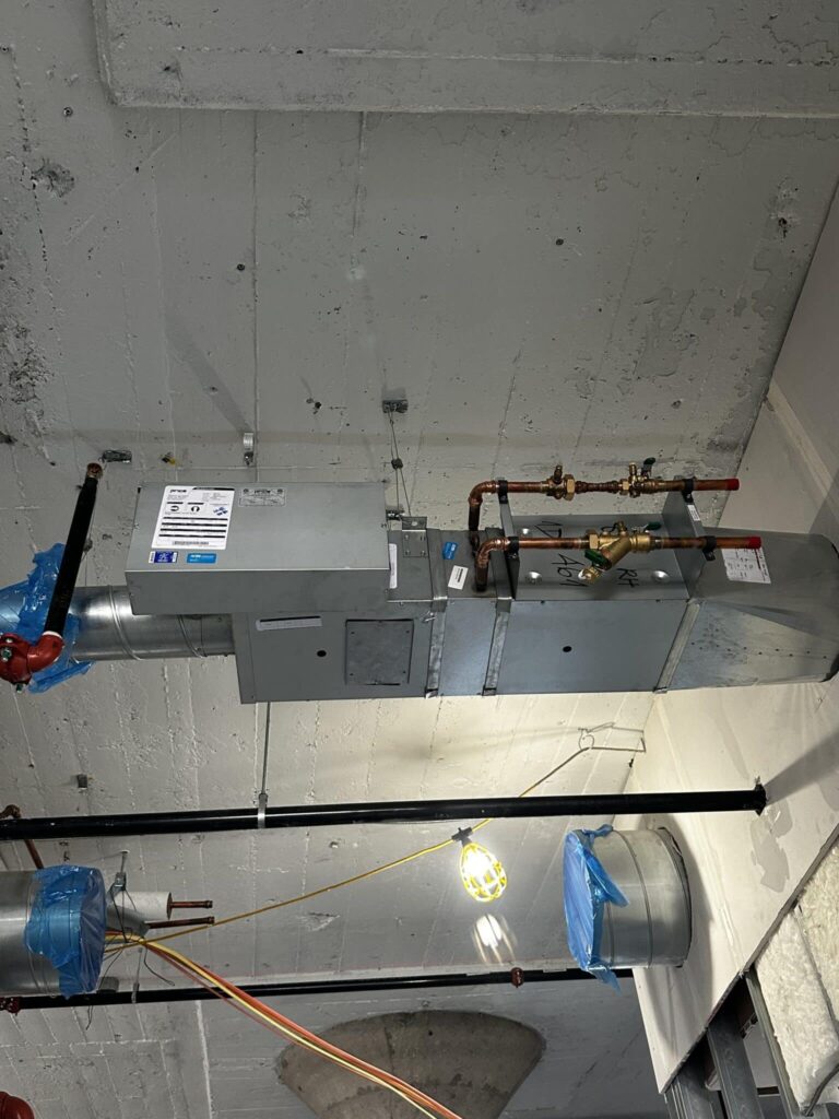 A view of an industrial ceiling featuring a large ventilation system with metal ducts and pipes. The ducts are wrapped with blue plastic, and signage is visible on one of the metal boxes. Adjacent pipes and a bright overhead light fixture are also visible, highlighting the textured concrete ceiling.