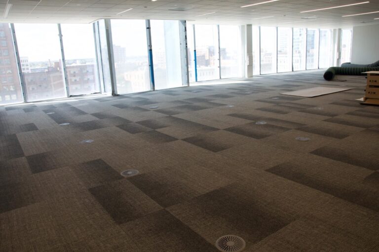A spacious, empty office room with large windows allowing natural light to flood in. The floor is covered with grey carpet tiles, and there are visible floor outlets. The windows offer a view of neighboring buildings in the background. The room is devoid of furniture, and there are some construction materials and a duct in the far corner, indicating ongoing setup or renovation work.