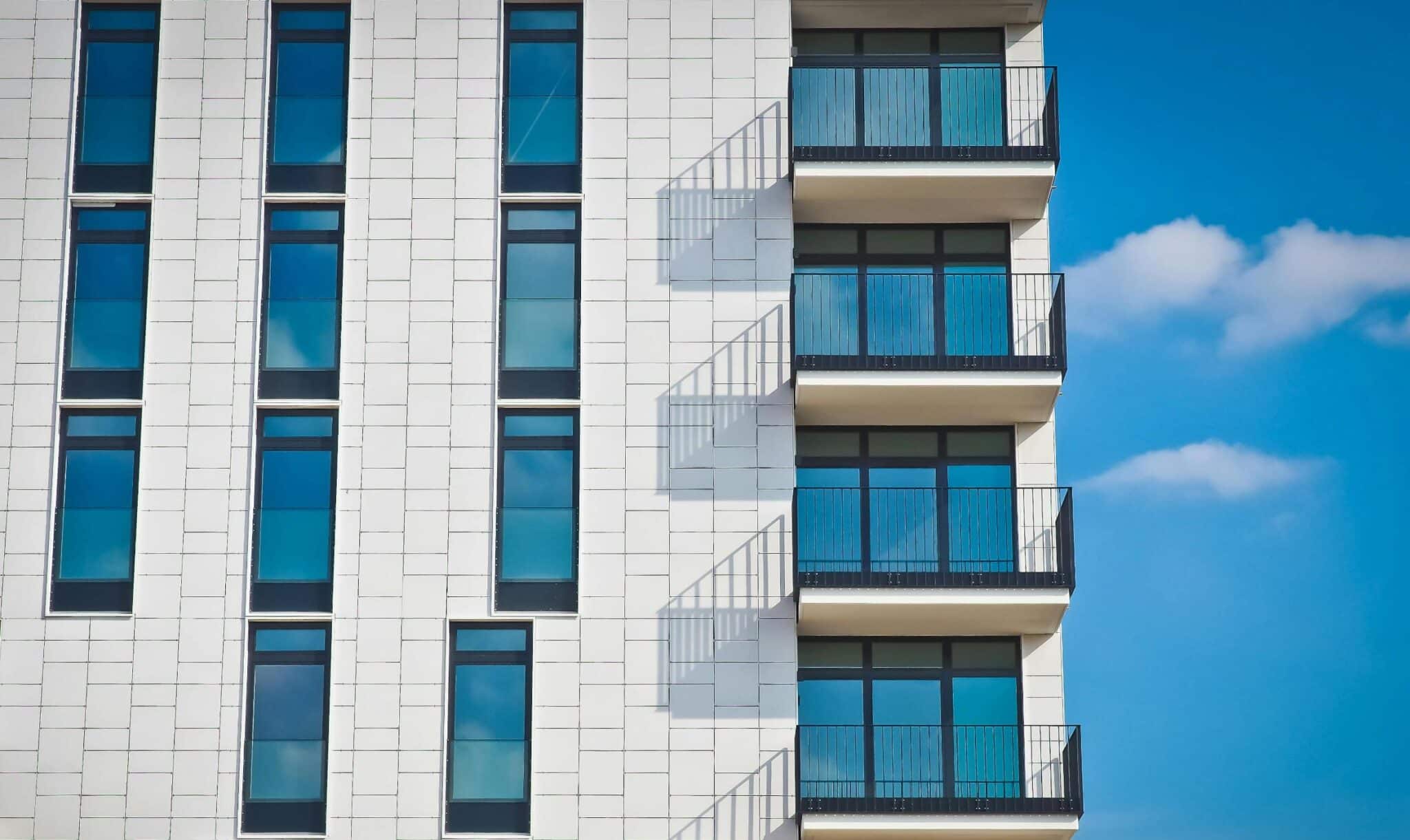 A modern apartment building with a white facade and rows of large, rectangular windows. The right side features several balconies with metal railings. The sky is clear with a few scattered clouds, adding a vibrant backdrop.
