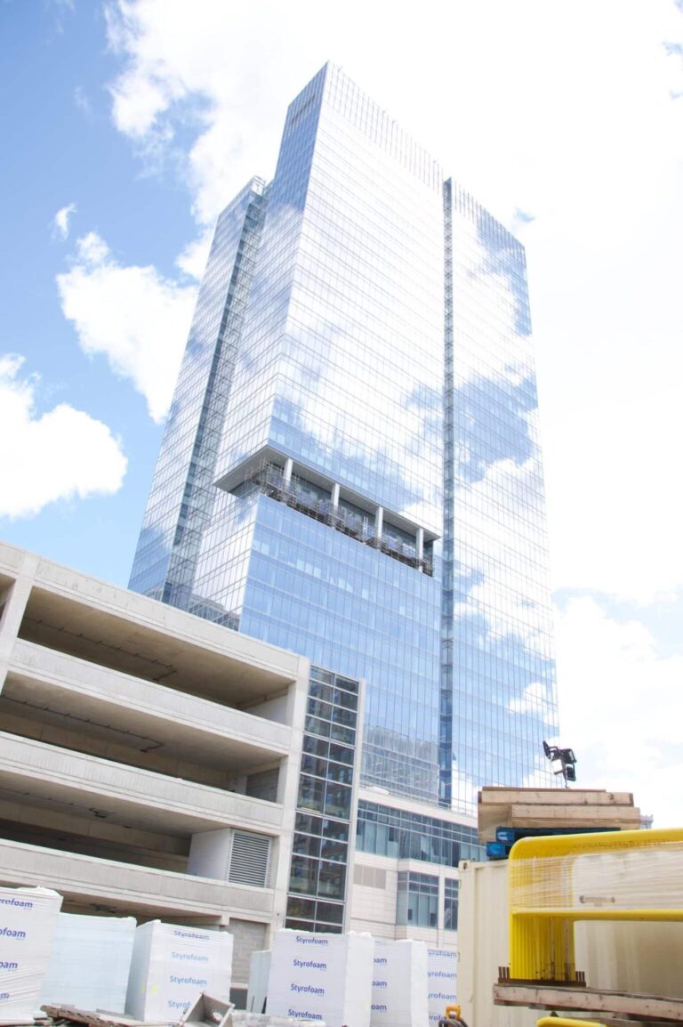 A tall modern glass skyscraper reflecting clouds stands near a partially built concrete structure. Styrofoam packaging and construction materials are visible in the foreground against a backdrop of a clear blue sky.