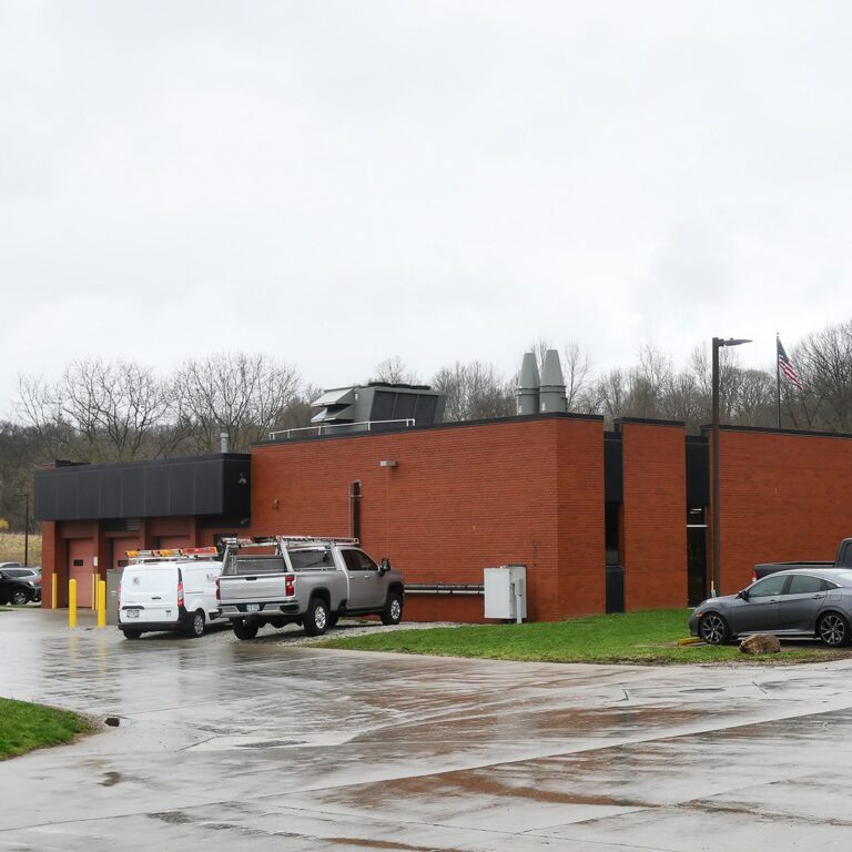 A modern industrial building with a red brick exterior and flat roof, featuring large vent structures on top. Several vehicles, including a white van and a silver truck with ladders on top, are parked on the wet concrete driveway outside. The sky is overcast and a tall flagpole with an American flag is visible next to the building.