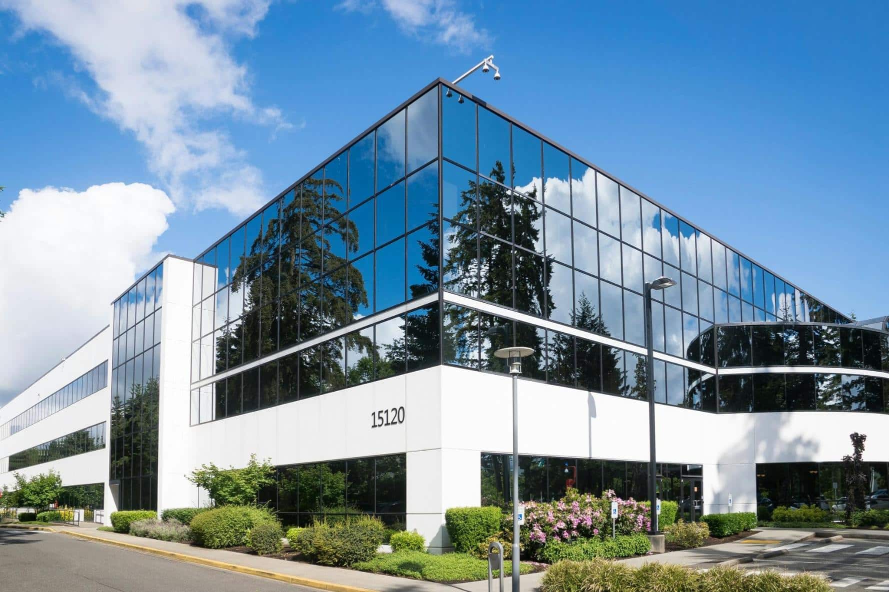 A modern office building with large glass windows reflecting the surrounding trees and blue sky, featuring a bright white facade with the number 15120. The foreground has neatly maintained shrubs and a small garden area, with two street lamps visible along the pathway.