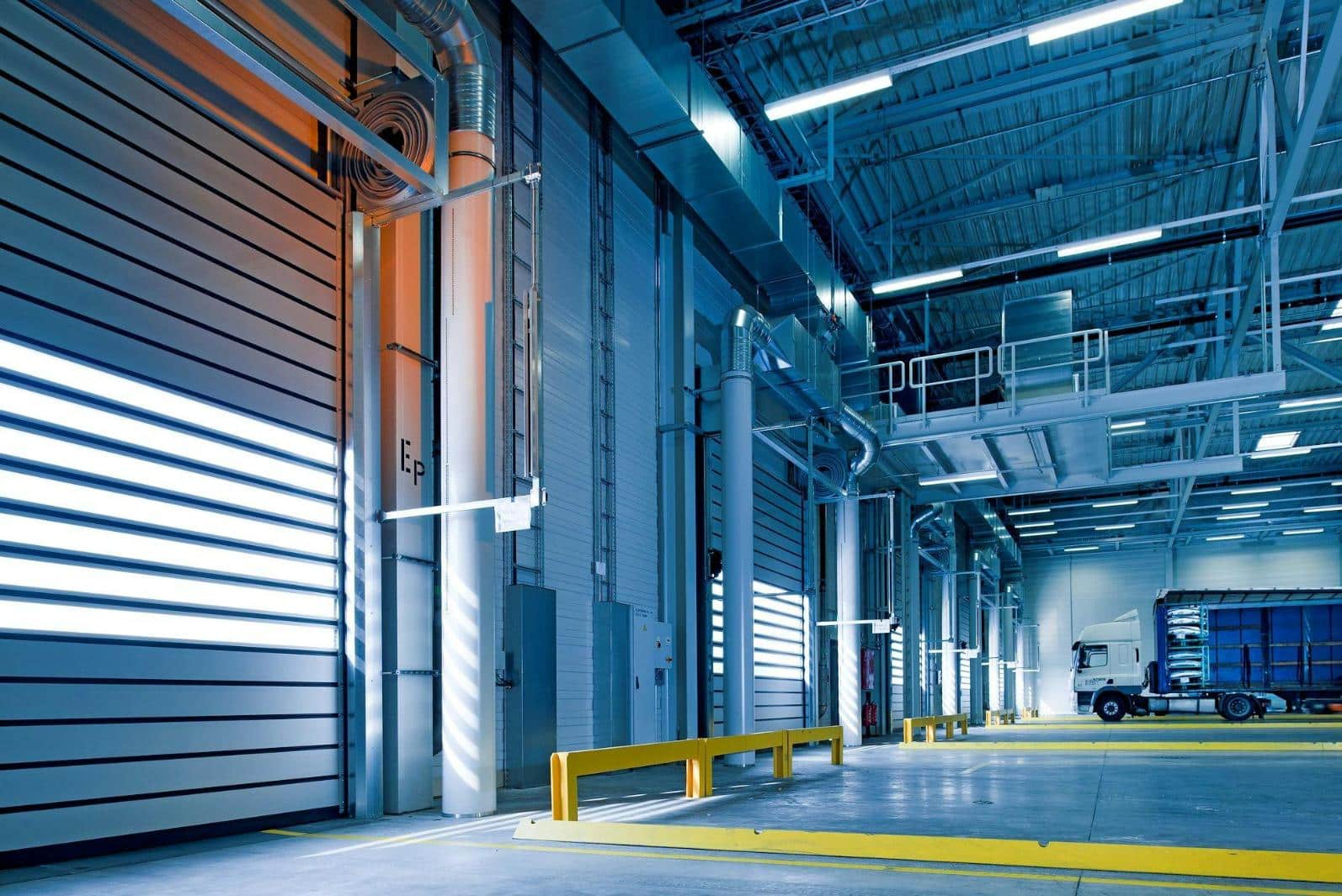 Interior of a modern warehouse with high ceilings, industrial lighting, and a white truck parked next to a loading dock. The space has metallic walls and bright yellow safety barriers along the floor.