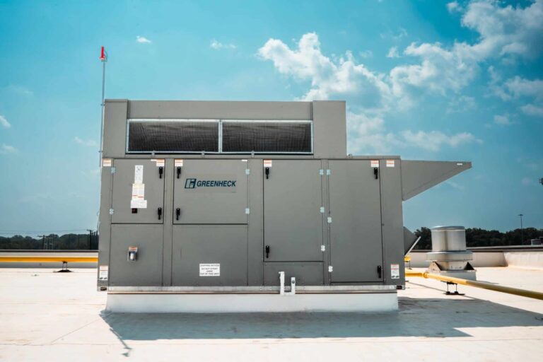 A Greenheck HVAC system installed on a rooftop against a blue sky with clouds. The unit is large, metal, and rectangular, with multiple access panels and a small red flag on a pole. The rooftop is expansive and flat, with a yellow pipe visible in the background and a silver ventilation cap nearby.