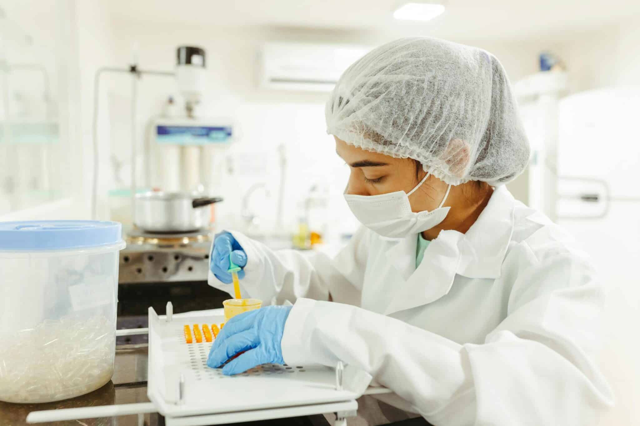 A scientist wearing a lab coat, mask, and hairnet is conducting a laboratory experiment. They are using a pipette to add liquid into a yellow container placed on a white tray. The scientist is focused on their task, and the laboratory equipment is visible in the background.