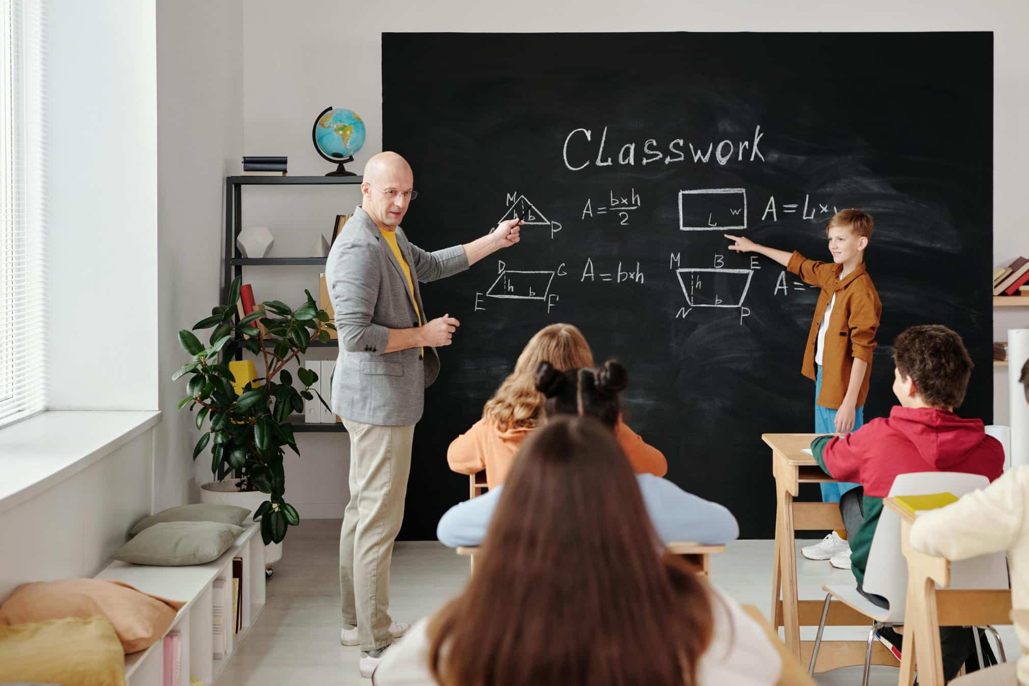 A classroom setting where a teacher and a student are at the front of the room. The teacher, wearing a gray blazer, points to a triangle with math formulas on a blackboard. The student, in a brown shirt, points to a rectangle on the blackboard. Several students are seated at desks, facing the blackboard. A globe and books are on a shelf nearby, with a chalkboard displaying geometry diagrams and the word 'Classwork' written on it.