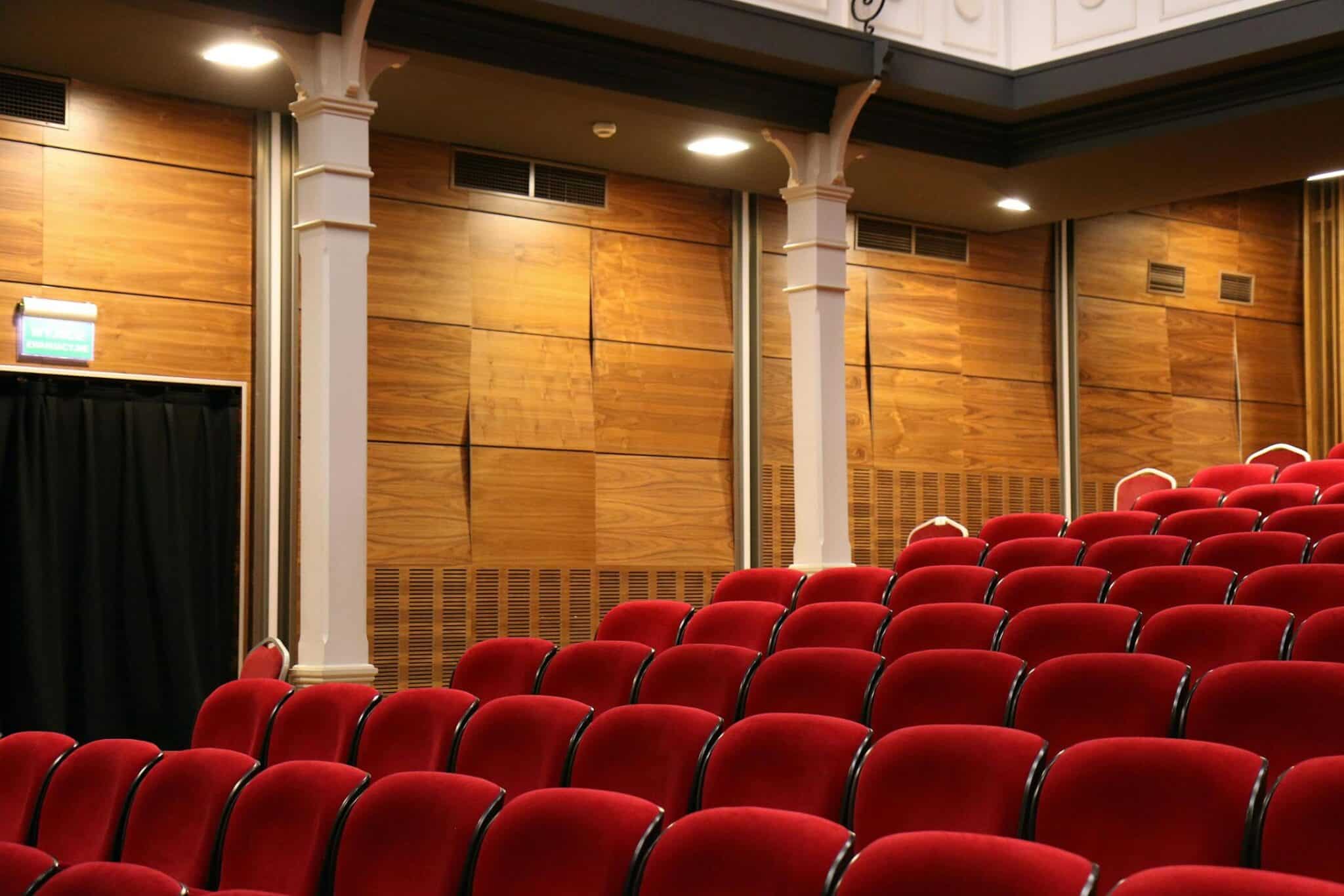 Rows of plush red theater seats in a modern auditorium, set against a backdrop of wooden-paneled walls and white columns, with ceiling lights softly illuminating the space.