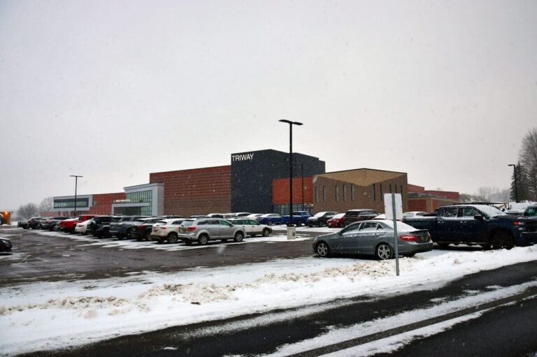 A large, modern building with the sign 'TRIWAY' visible on one of its walls, surrounded by a busy parking lot filled with cars. The ground and rooftops are covered with a light layer of snow, indicating a wintry setting. The sky is overcast, and there are streetlights and a few trees in the background.